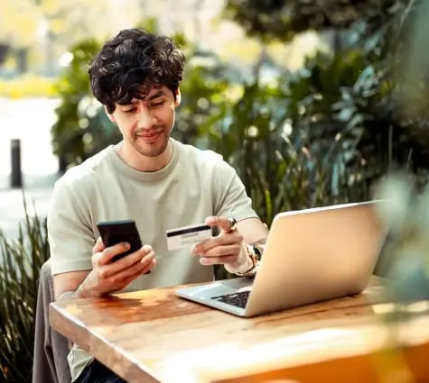 Young Latino or Hispanic man sitting at a table outside with laptop and looking at phone and credit card