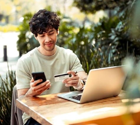 Young Latino or Hispanic man sitting at a table outside with laptop and looking at phone and credit card