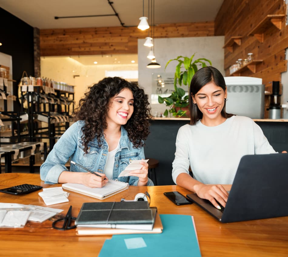 Two women log receipts using a laptop, notebook, and calculator.