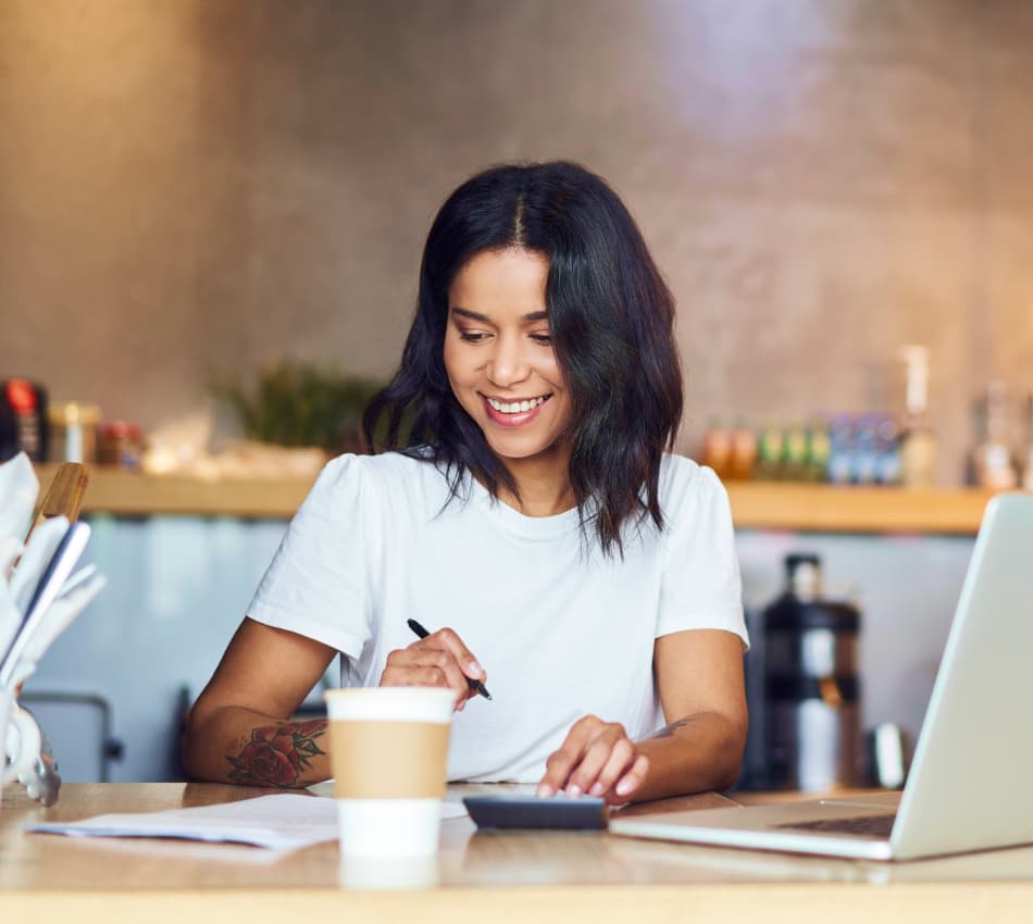 Woman in coffee shop aligns numbers on paper and laptop using calculator.