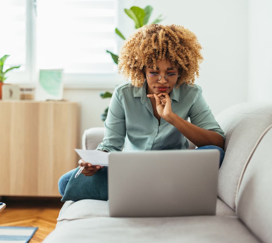 Woman reads from laptop screen while holding paper and a pen, sitting on couch.