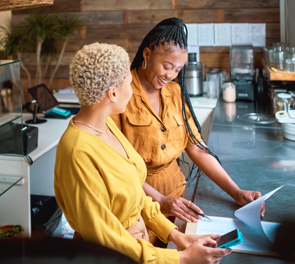 Two women look over a clipboard behind the register at a shop.
