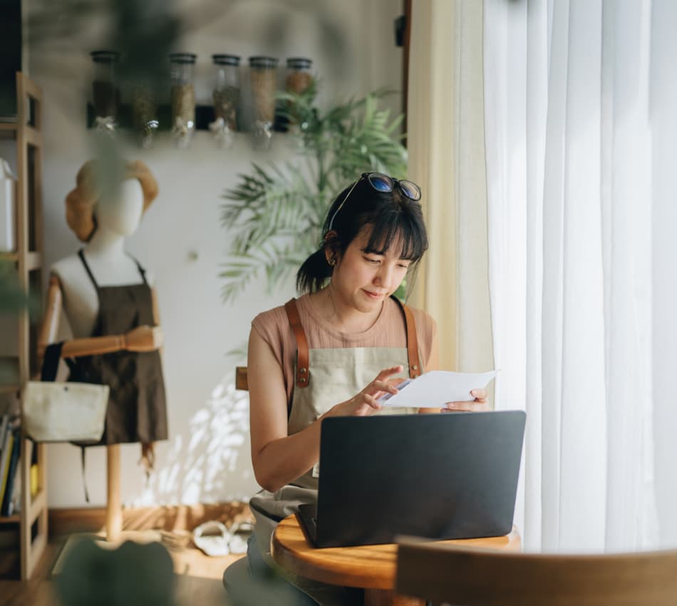 Female shop owner reviews piece of paper while sitting in front of her laptop.