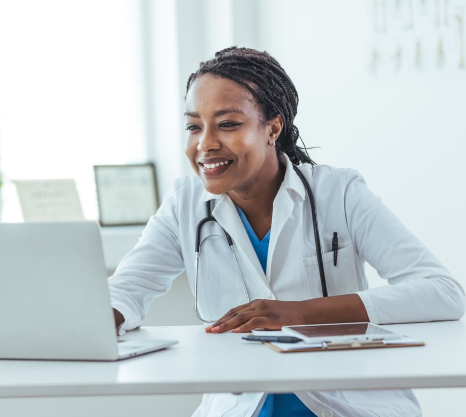 A female doctor sits at her desk and looks at her laptop while smiling.