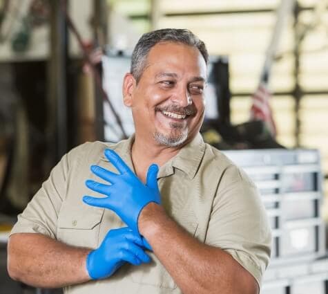 Middle-aged Hispanic man smiling while putting on rubber gloves in his auto repair shop.