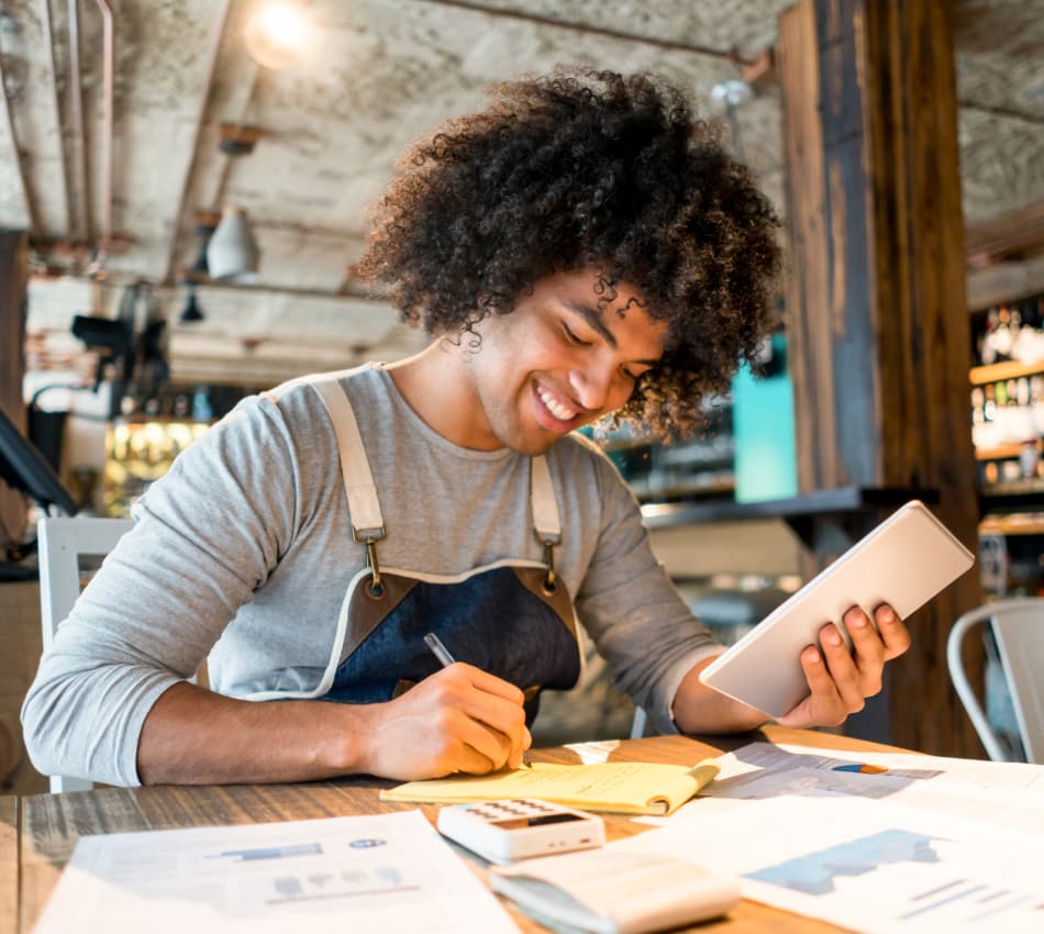 Liquor store owner reconciling his finances with a tablet, notepad, and calculator.