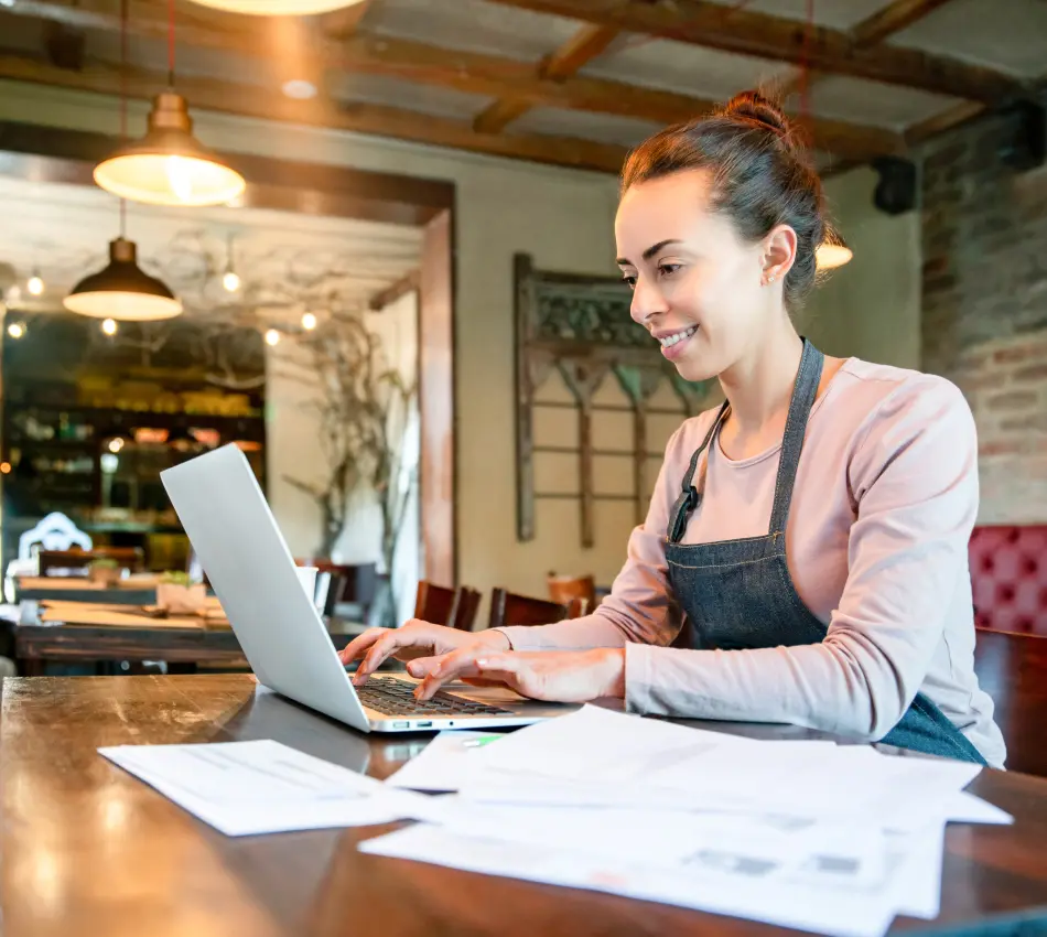 Female business owner types on laptop next to stack of papers at her cafe.
