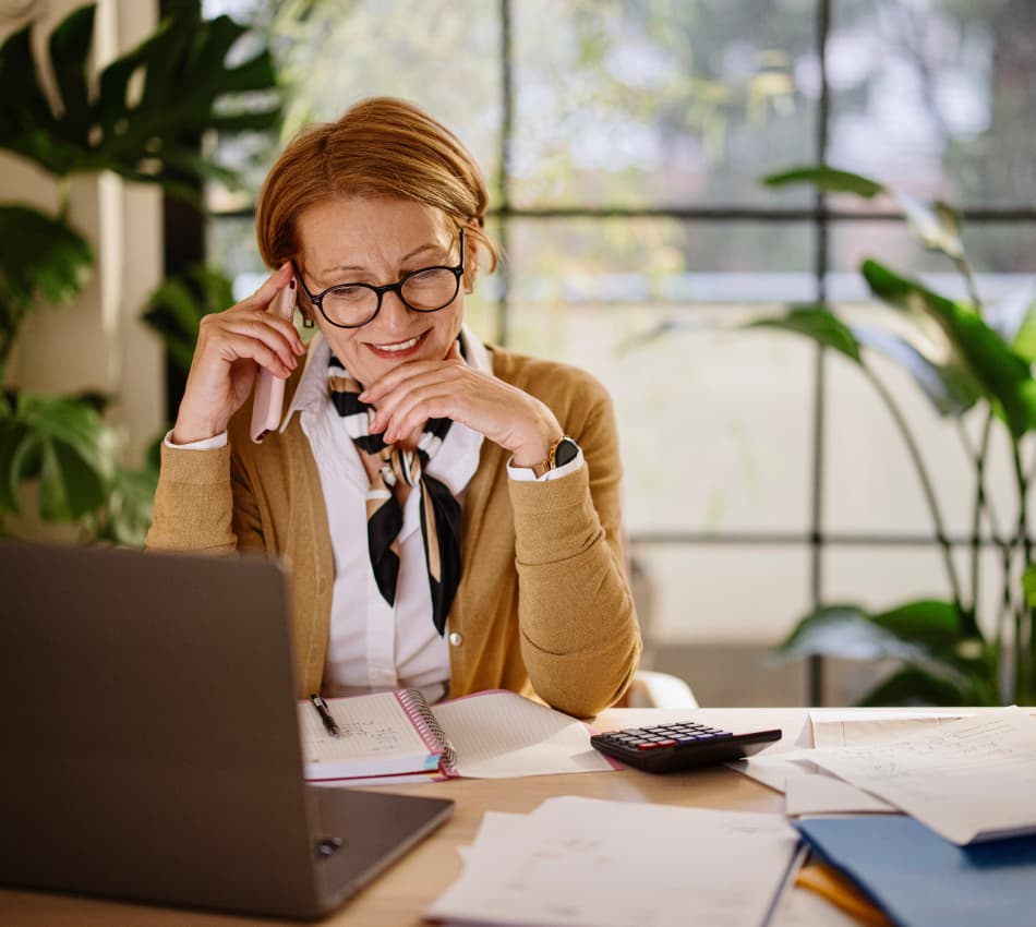 Woman on a phone call, sitting at laptop with notebook and calculator nearby.