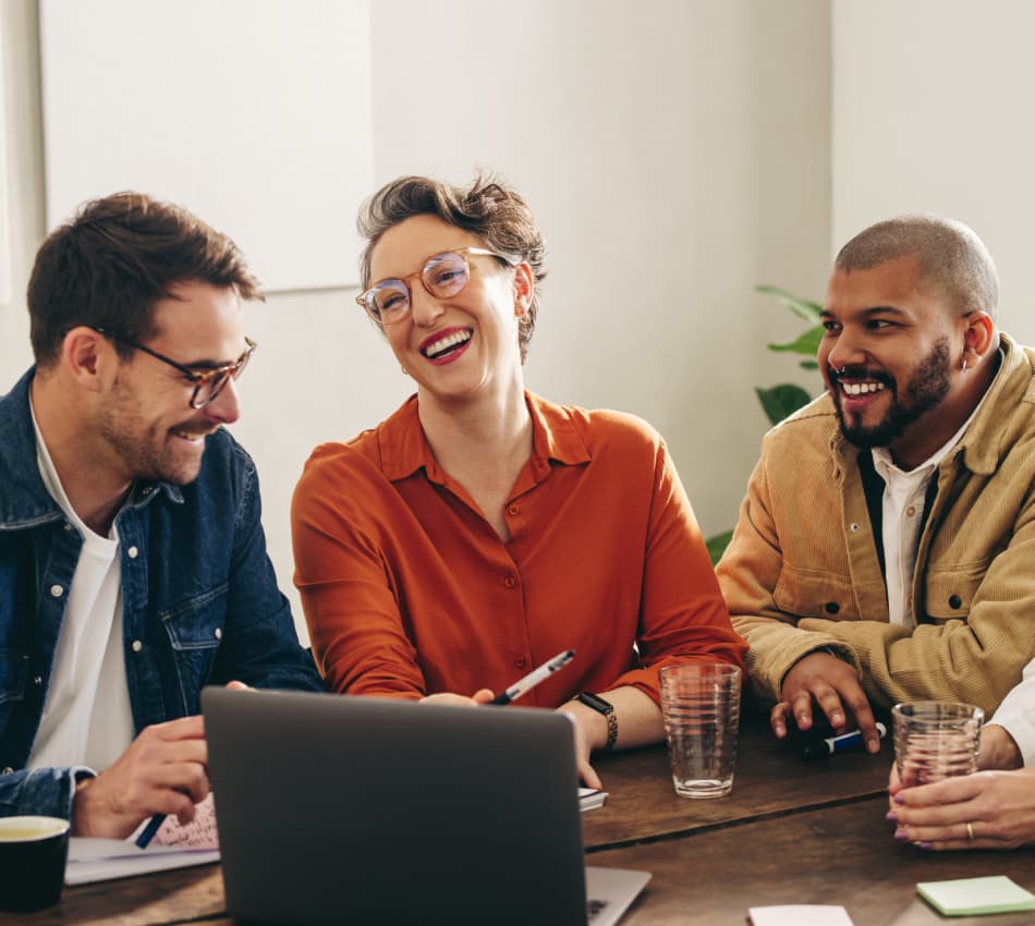 A group of employees laugh in front of a laptop while sitting at a table.