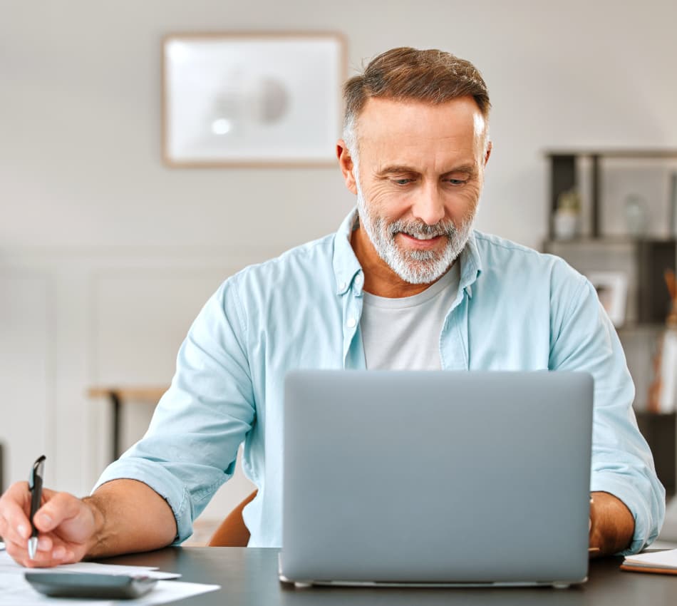 Male business owner smiles at laptop while writing on a piece of paper next to his calculator.