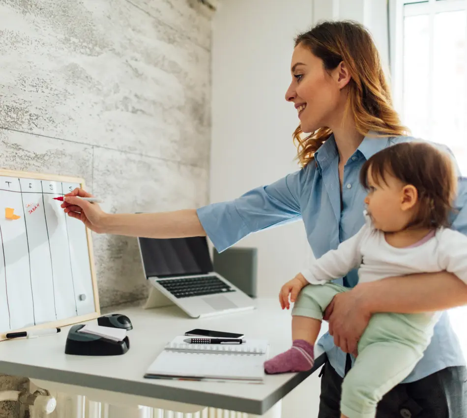 A female small business owner writes out her schedule on a whiteboard while holding her baby near her desk.