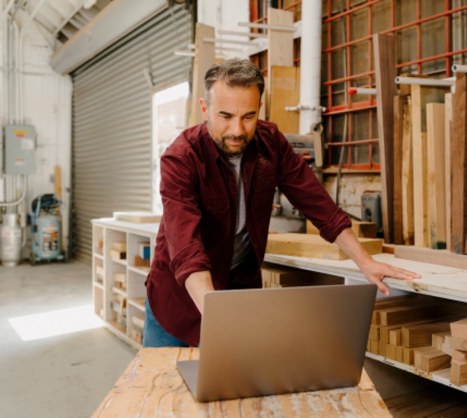 A construction business owner standing in a warehouse, surrounded by wood and tools, looking at his laptop.