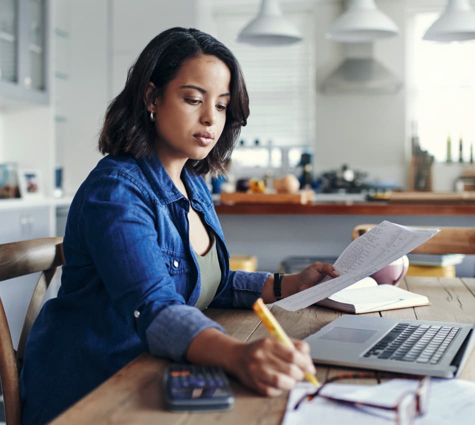 Female business owner crunches numbers at her kitchen table with an expense sheet, calculator, and laptop.
