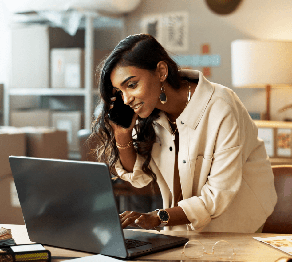 Ecommerce business owner manages her inventory from her laptop while on a phone call, with boxes in the background.